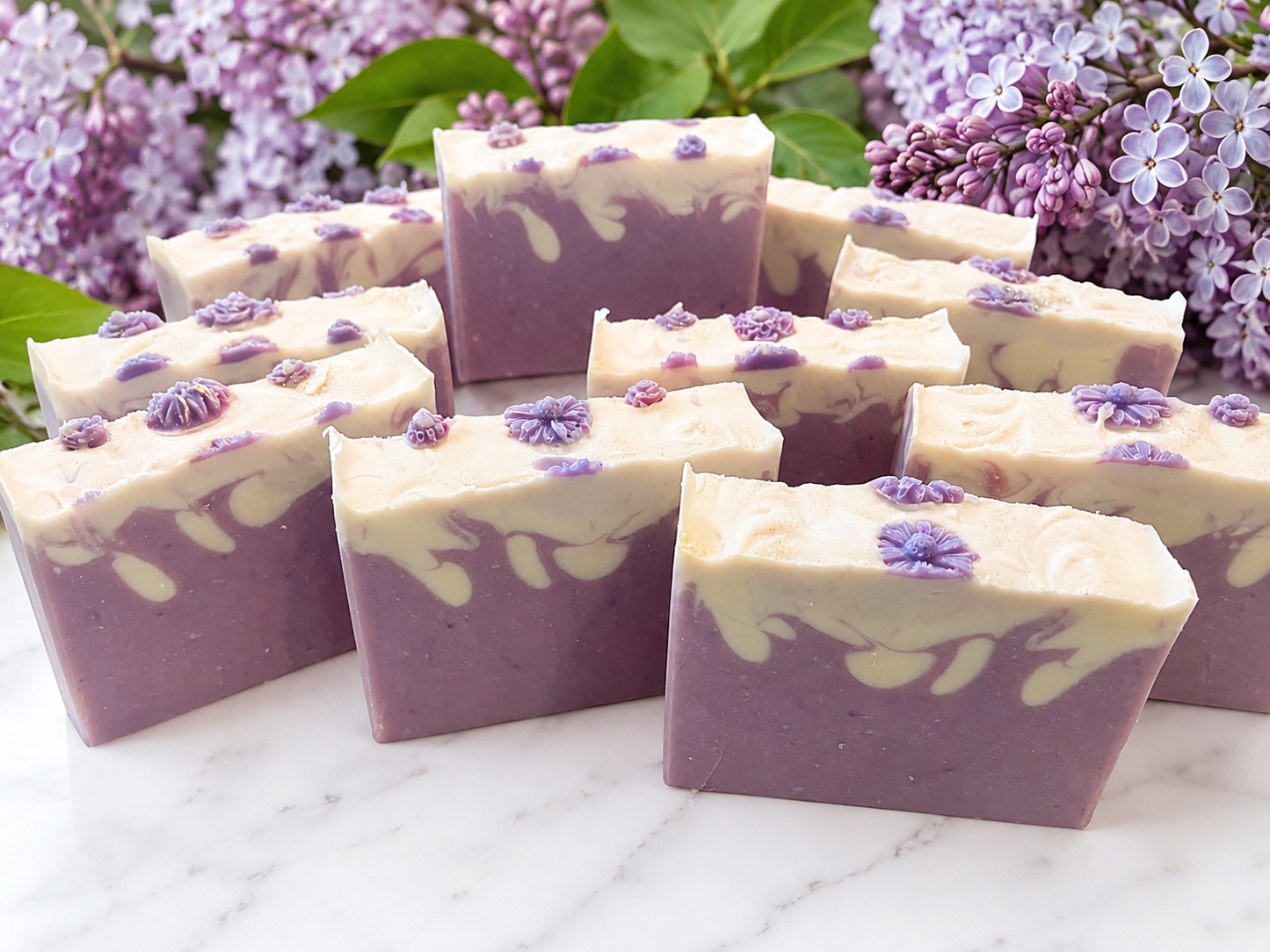 Several rectangular bars of lavender soap with cream swirls and small purple flower decorations, arranged on a white marble surface with lilac flowers in the background.